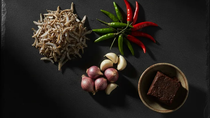 Ilustrasi dijana AI — bahan sambal ikan bilis disusun di atas permukaan batu gelap: timbunan ikan bilis kering, cili hijau dan cili merah, bawang merah, bawang putih dan sedikit belacan dalam mangkuk kecil