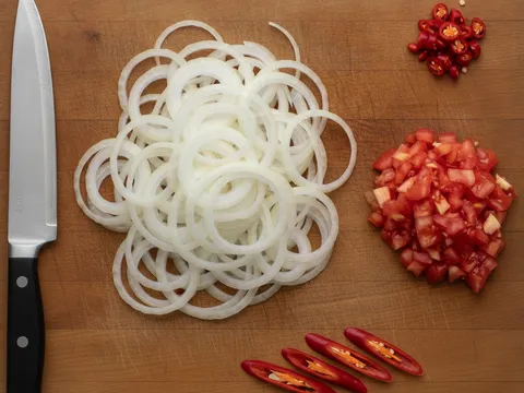 Bawang besar hiris nipis, tomato dadu, cili padi dan cili merah dihiris di atas papan pemotong