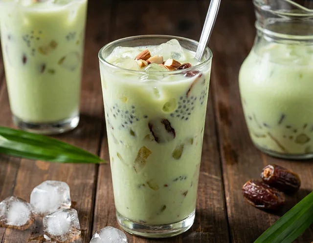 Jag kaca air katira hijau lembut bersebelahan dua gelas air katira berais, dengan daun pandan, kurma dan badam cincang di atas meja kayu