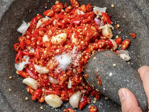 Cili padi merah dan bawang putih ditumbuk kasar dalam lesung batu menjadi sambal geprek mentah
