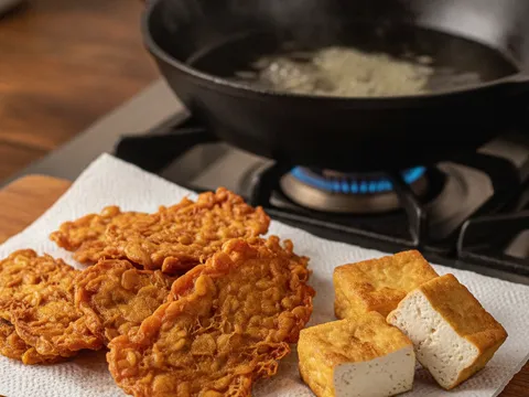 Kepingan tempe dan tauhu goreng keemasan ditoskan di atas kertas serap minyak di sebelah kuali berminyak panas