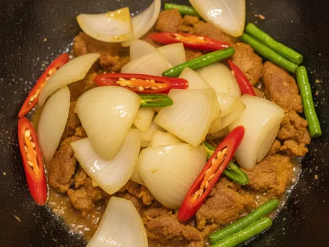 Hirisan bawang besar, cili merah dan cili padi hijau dimasukkan ke dalam kuali daging goreng kunyit