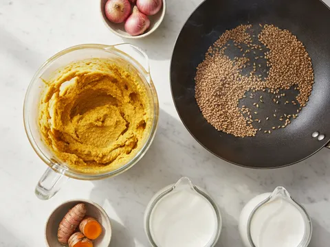 Bahan kisar gulai ayam berwarna kuning oren dalam blender di sebelah kuali kering berisi biji ketumbar dan jintan yang disangai