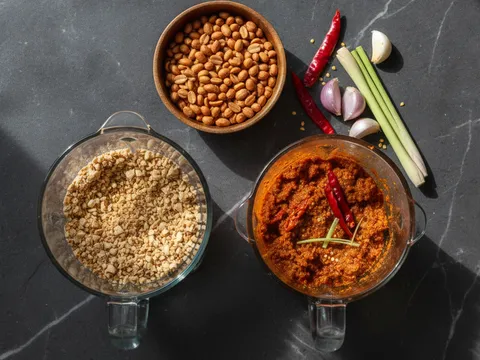 Kacang tanah sangai dikisar kasar dalam blender — tekstur serpihan kasar, bukan halus seperti butter; pes rempah merah-oren dari cili kering dan rempah dalam mangkuk berasingan