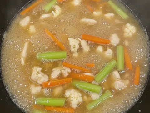 Stok ayam mendidih dalam kuali bersama hirisan lobak merah, kuntum bunga kobis dan batang sawi