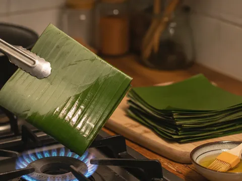 Kepingan daun pisang dilayur di atas api dapur sehingga hijau berkilat dan lembut