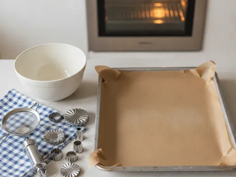Dulang pembakar dilapik kertas baking di atas meja dapur, di sebelahnya acuan semperit dan mangkuk adunan