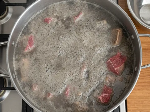 Tulang dan daging lembu dalam periuk besar berisi air rebusan keruh berbuih kelabu di permukaan