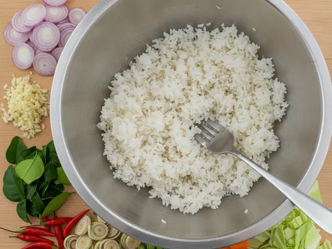 Nasi putih sejuk dileraikan dengan garfu dalam mangkuk besar, dikelilingi bahan potong seperti bawang merah, serai dan kacang panjang