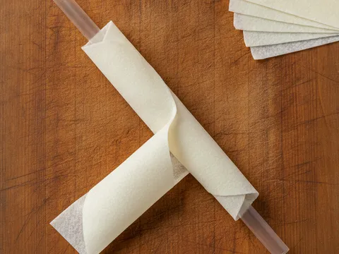 Sehelai kulit popia sedang digulung memanjang di atas straw plastik di atas papan pemotong